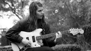 Alex Lahey sitting down and playing her guitar.